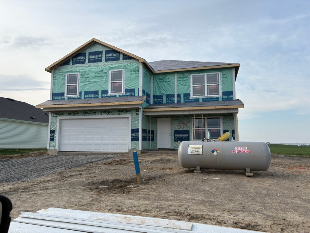 Newly constructed house by DR Horton, exterior wrapped in insulation with a garage and propane tank visible in front.