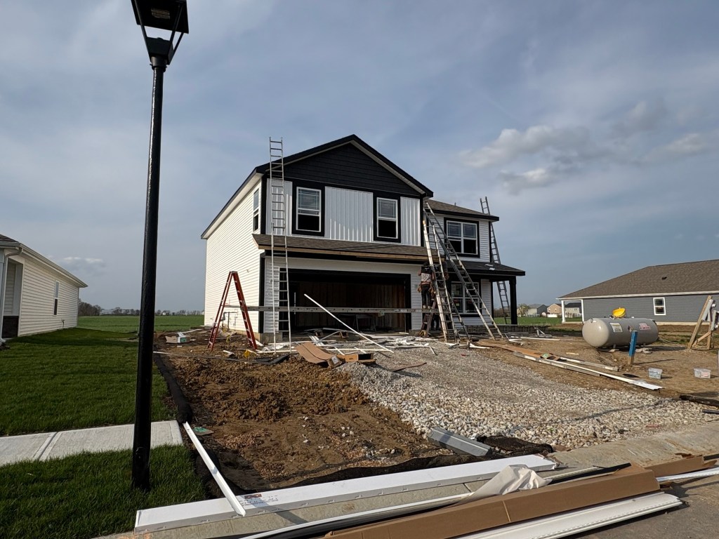 A newly built house featuring a black and white farmhouse design, with construction workers on ladders working on the exterior. The surrounding area is partially landscaped with grass and gravel.