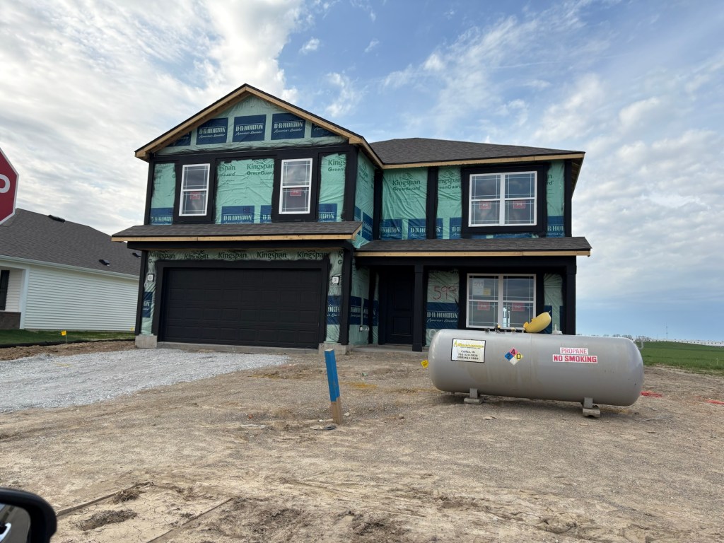 A newly constructed two-story house with green insulation and black accents, located on a gravel lot. There is a propane tank in front and a stop sign in the background.