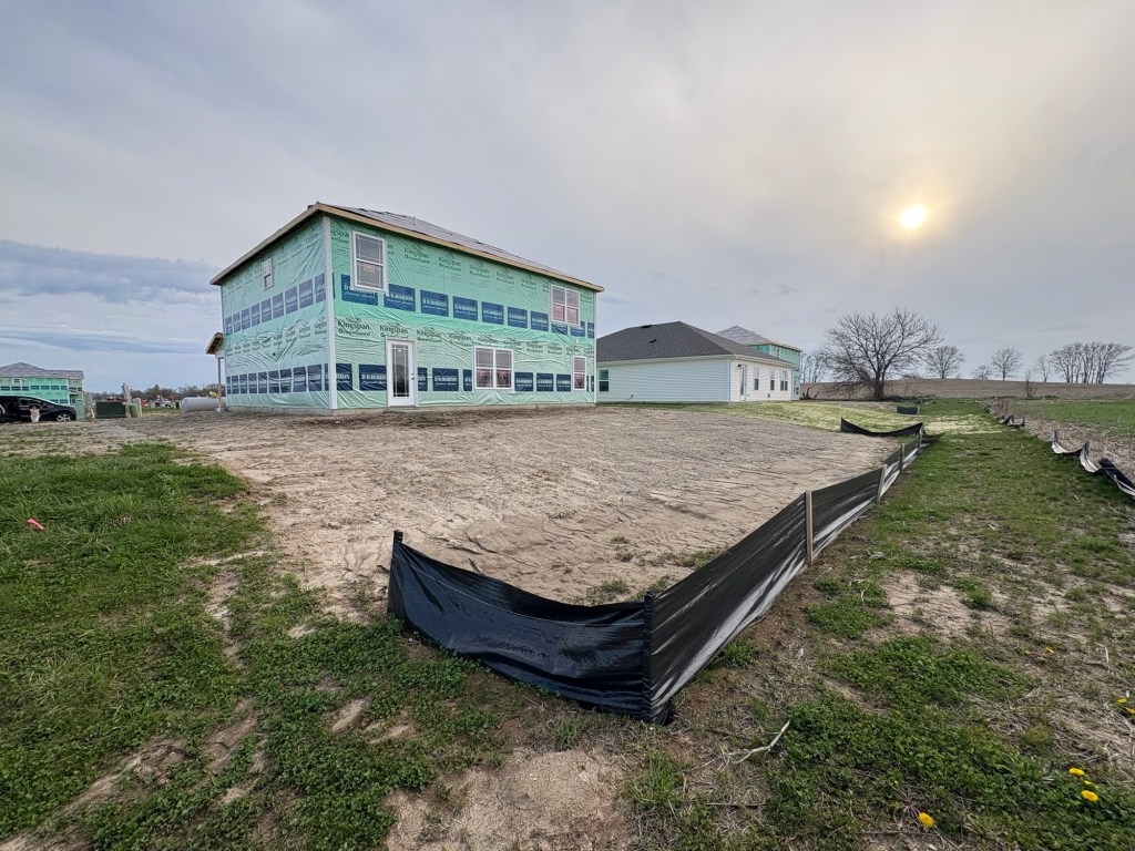 A newly constructed house in the building phase, surrounded by a fenced yard and other houses in the background under a cloudy sky.